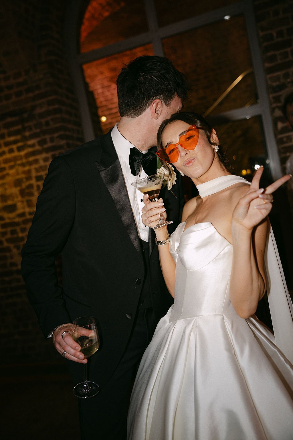 A couple dressed in formal attire at a party, the man in a black tuxedo with a bow tie and the woman in a white satin gown with heart-shaped orange sunglasses, holding drinks, making peace signs, and smiling.