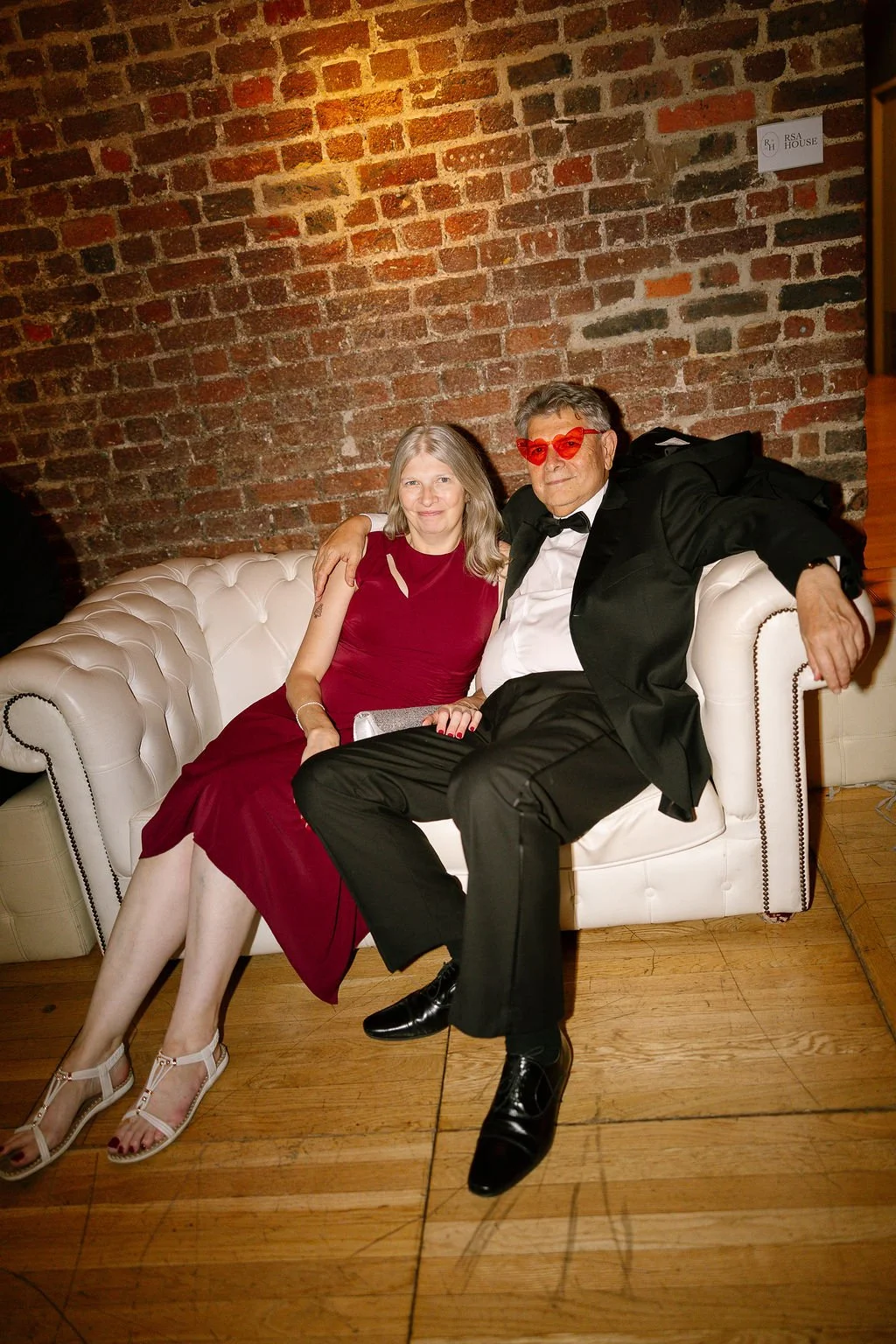 A woman in a red dress and a man in a black tuxedo with red heart-shaped sunglasses sitting together on a white couch against a brick wall.