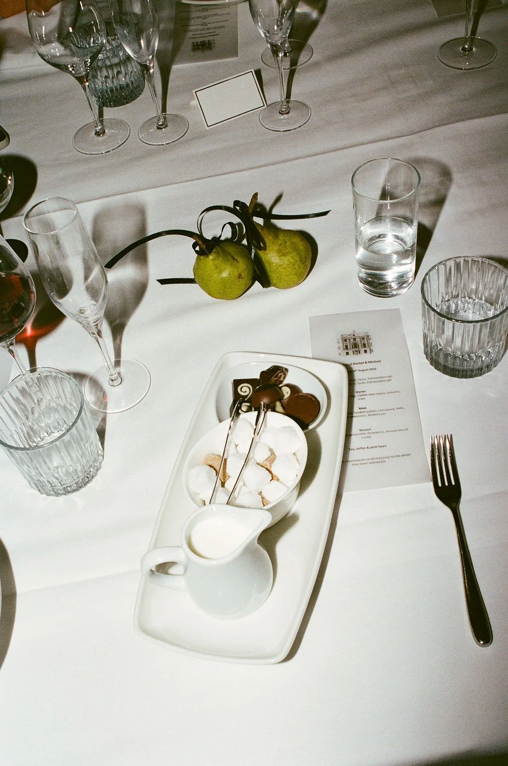 A table setting with a white plate of assorted chocolates, a small pitcher of cream, and a bowl of marshmallows. There are also pears attached to a branch, several glasses of water, and wine glasses. A printed menu is partially visible on the table.