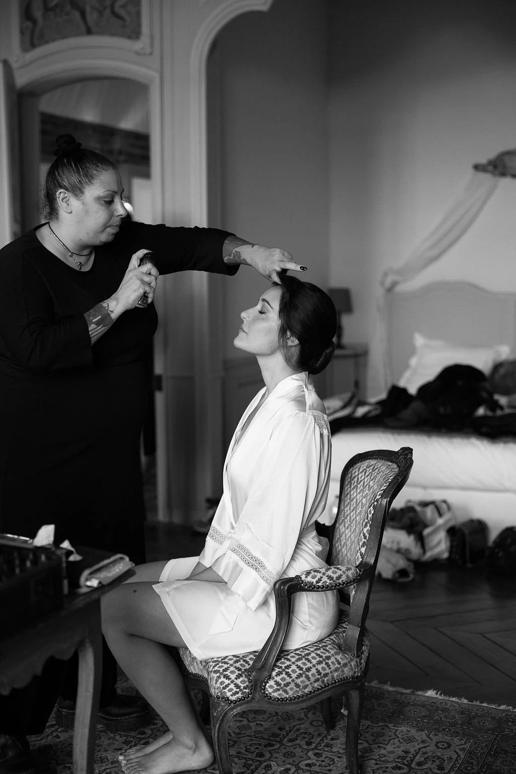 A woman in a silk robe is sitting with eyes closed as a makeup artist applies makeup to her face in a bedroom.