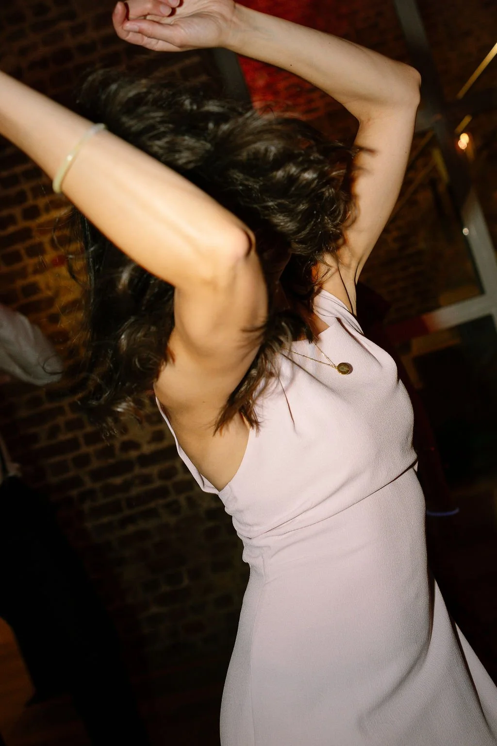 A woman with dark, curly hair is dancing with her arms raised above her head, wearing a light pink dress and a necklace, in a dimly lit indoor space with a brick wall background.
