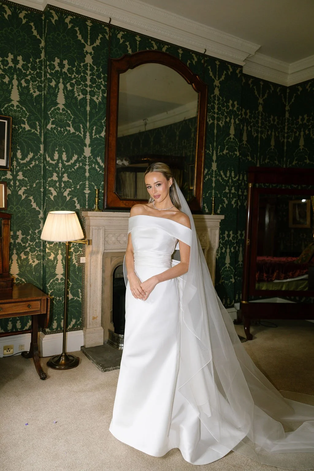 A bride in a white off-the-shoulder wedding gown with a long train, standing indoors in front of a fireplace with a large mirror. The room has dark green patterned wallpaper, a side table with a lamp, and antique furniture.