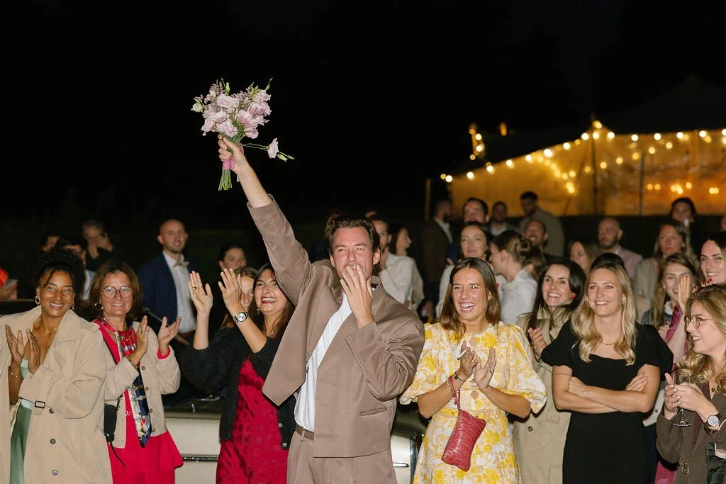 A groom in a beige suit holding a bouquet of flowers raises his hand and covers his mouth in surprise, surrounded by smiling guests at a wedding reception at night with string lights in the background.