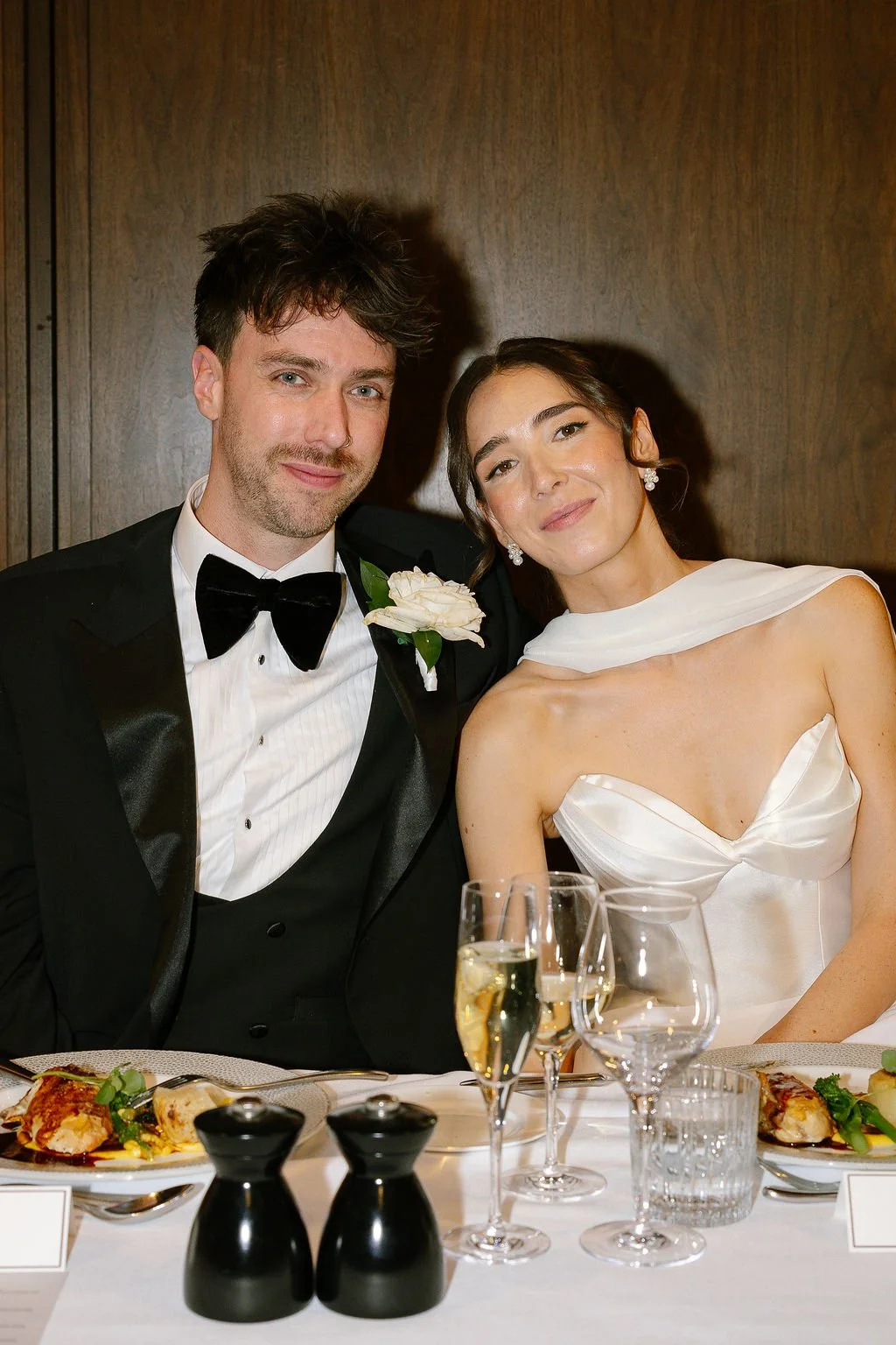 A newlywed couple in formal attire sits at a wedding reception table, smiling. The groom wears a black tuxedo with a bow tie and a white flower boutonniere, and the bride wears a strapless white satin dress with a draped white shawl. The table has el