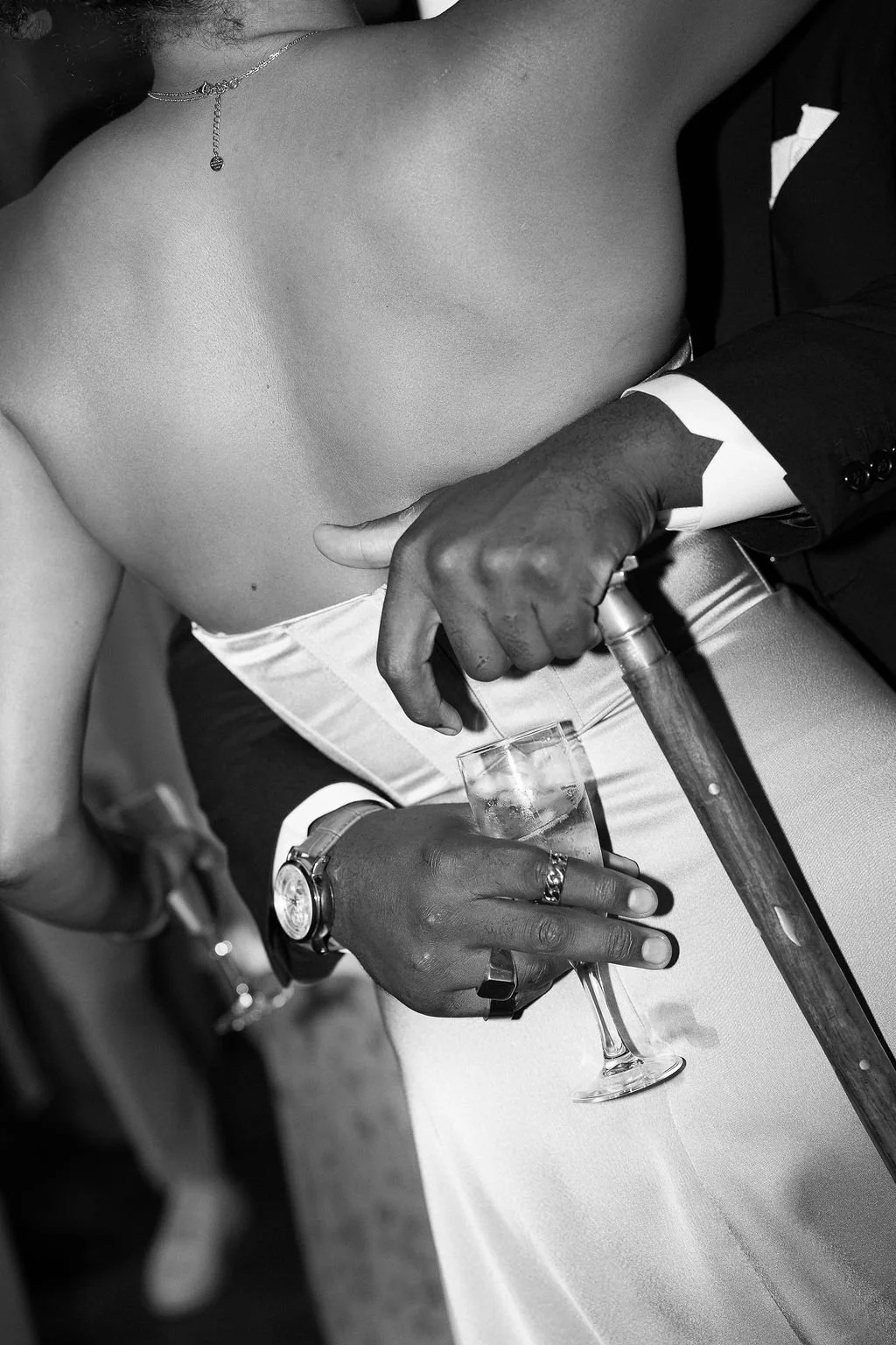 A black and white close-up photo of a person with bare shoulders, holding a glass of liquor, with another person wearing a suit helping them with a cane. The person holding the glass is wearing a watch and rings.