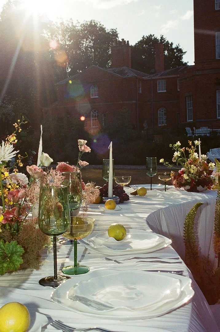 Elegant outdoor dinner table set with white plates, green and clear glassware, yellow lemons, and floral centerpieces, with a brick house and trees in the background during sunset.