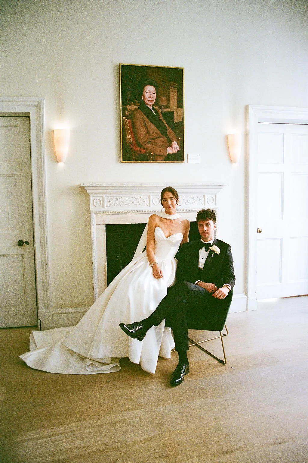 A bride in a white wedding gown and a groom in a black tuxedo sitting in front of a fireplace in a bright room, with a portrait of an older woman hanging on the wall above. The bride is standing with her hand on the groom's knee, and they are both looking at the camera.