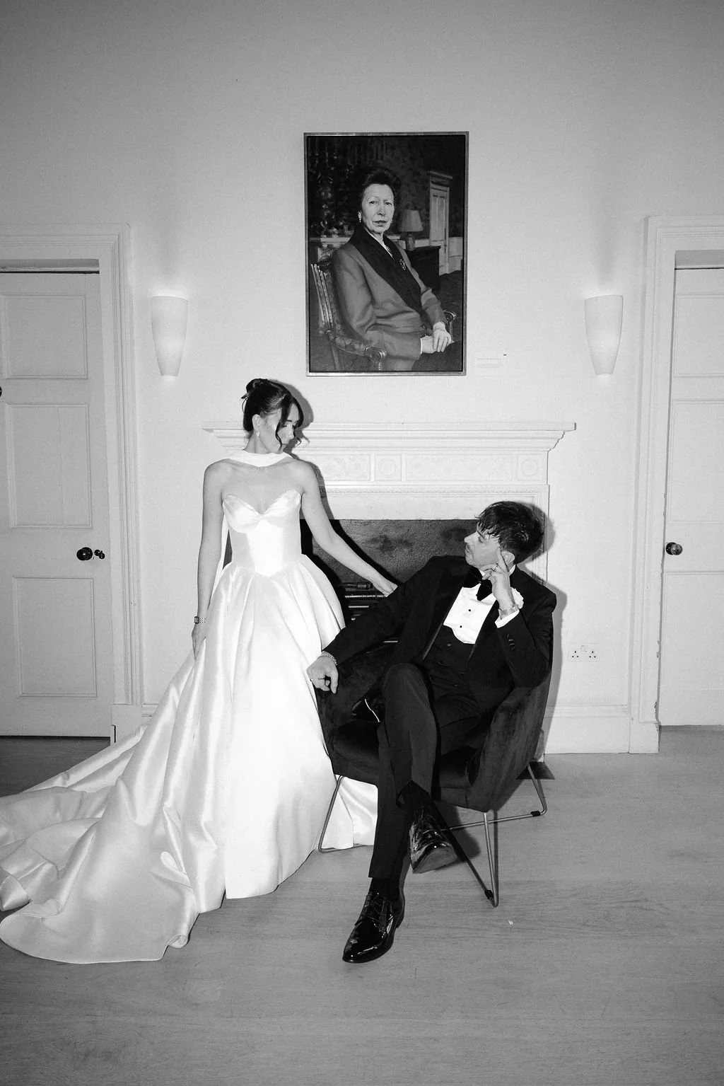 Black and white photo of a woman in a wedding dress and a man in a tuxedo inside a room, with a large portrait hanging above them. The woman is standing next to the man, who is sitting in a chair, looking up at her. The room has two closed doors and 