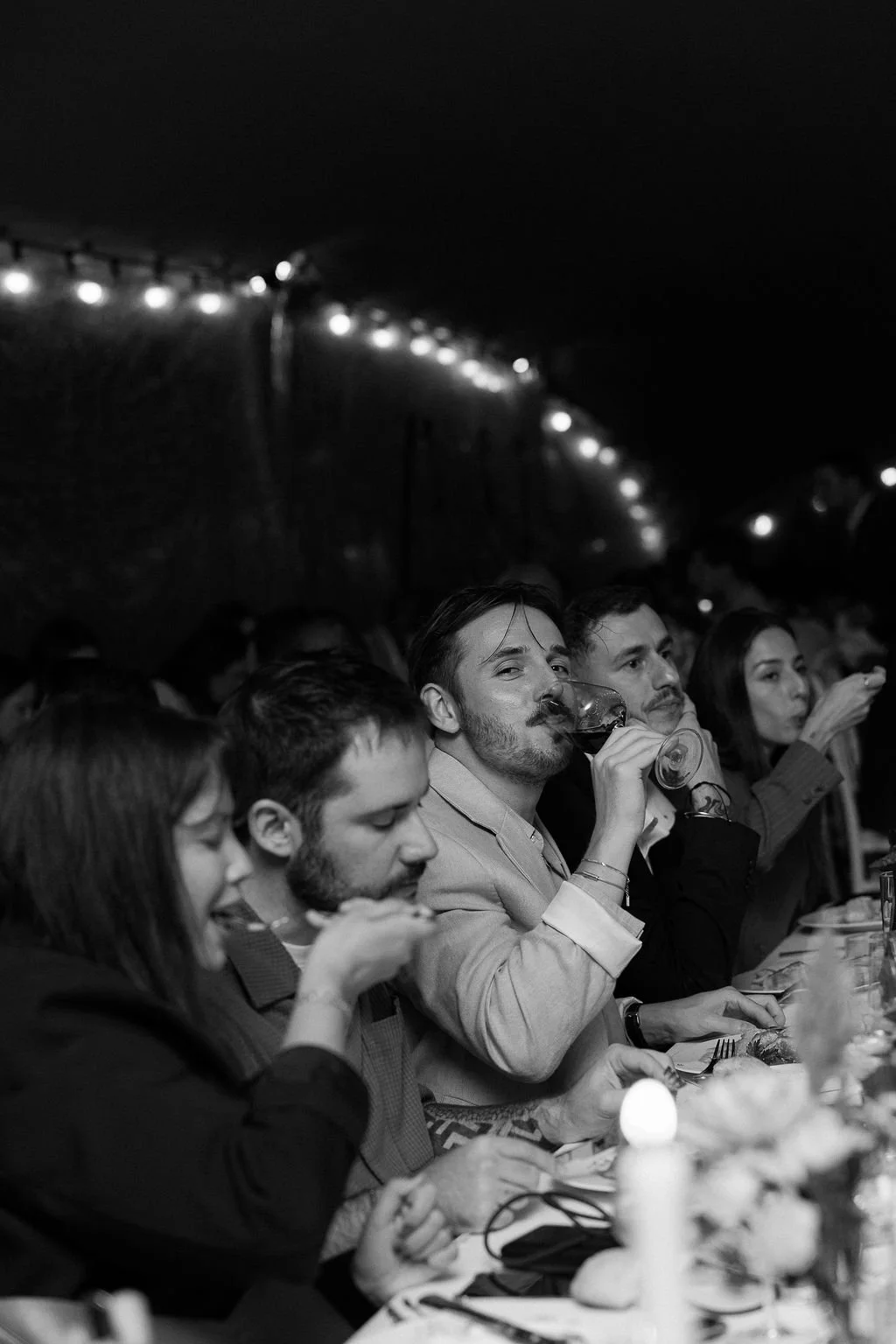 A group of people sitting at a long table enjoying a meal, with some drinking wine. The scene is indoors at night, with string lights overhead.