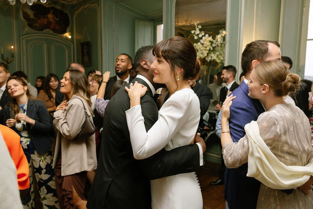 Couple dancing closely at a formal event with multiple other couples and guests, dressed in elegant attire, in a decorated, historic ballroom.