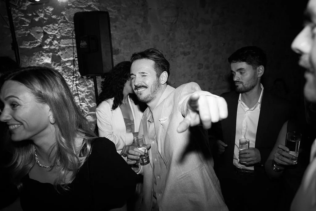 Group of people at a party, smiling and enjoying drinks, with one person pointing and laughing, in a dimly lit room with a stone wall background.