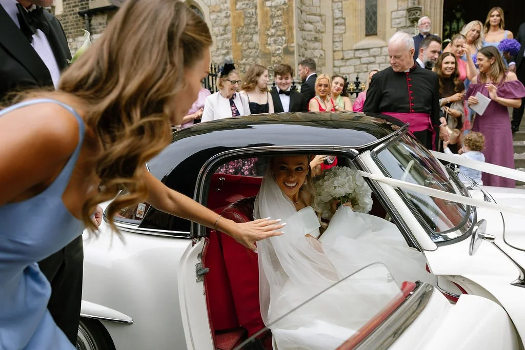 Bride smiling and holding flowers inside a vintage car at a wedding, with guests gathered outside a brick church in the background.