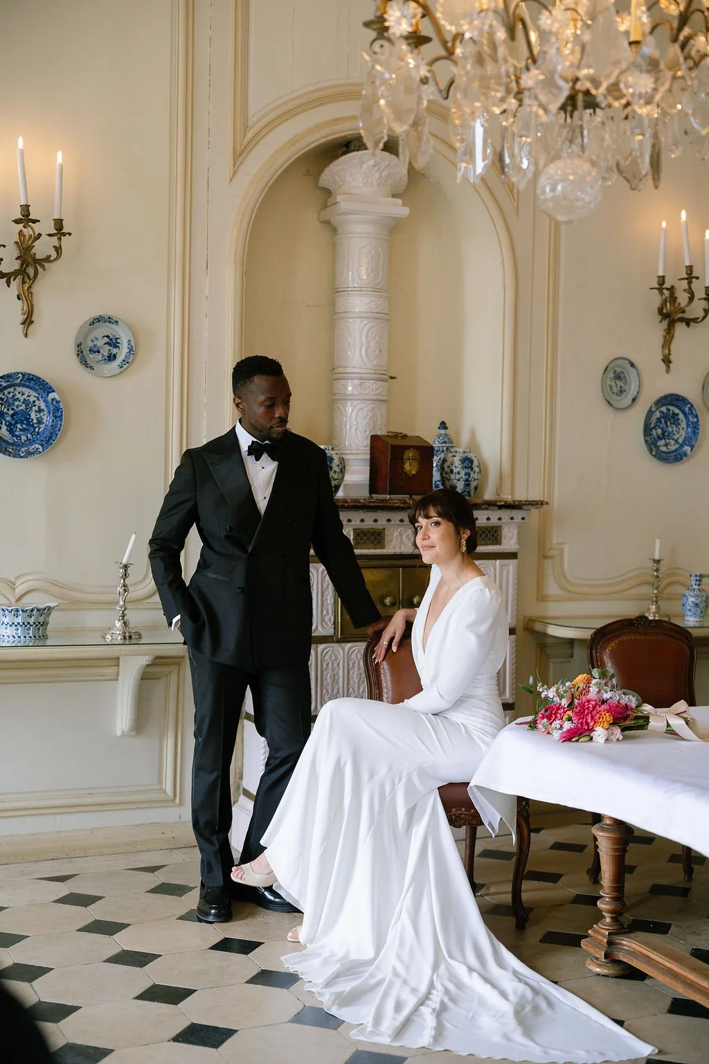 A man in a black tuxedo with a bow tie stands beside a seated woman in a white dress in an elegant dining room. The woman has dark hair and is sitting with her hand on her lap, looking at the man. The room features a chandelier, blue and white decora