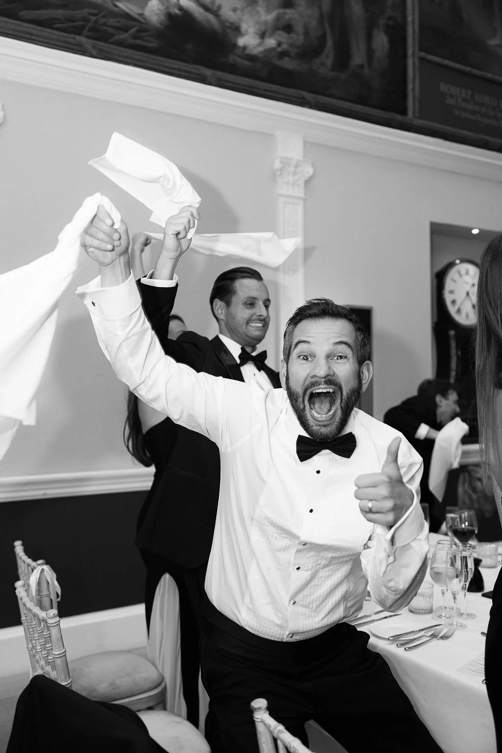 Men dressed in formal tuxedos celebrating at a dinner party, with one man in the foreground shouting and pointing, and others smiling and raising napkins.