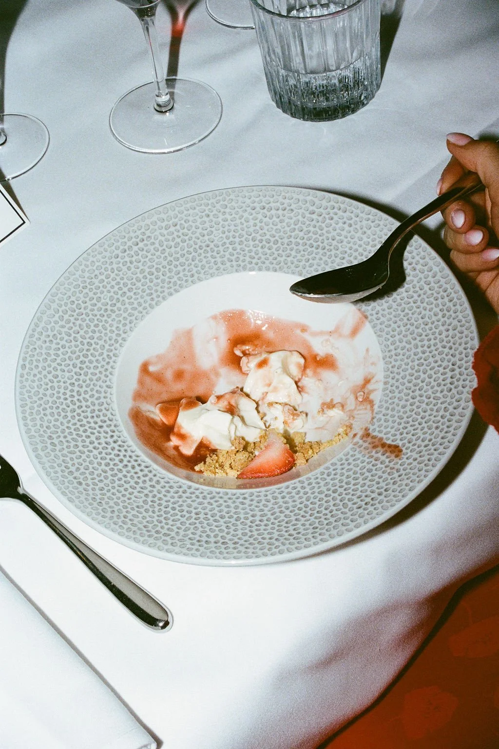 Empty dessert plate with strawberry slices, whipped cream, and crust remnants, held by a person's hand with a spoon, on a white table with wine glasses and water glass in the background.