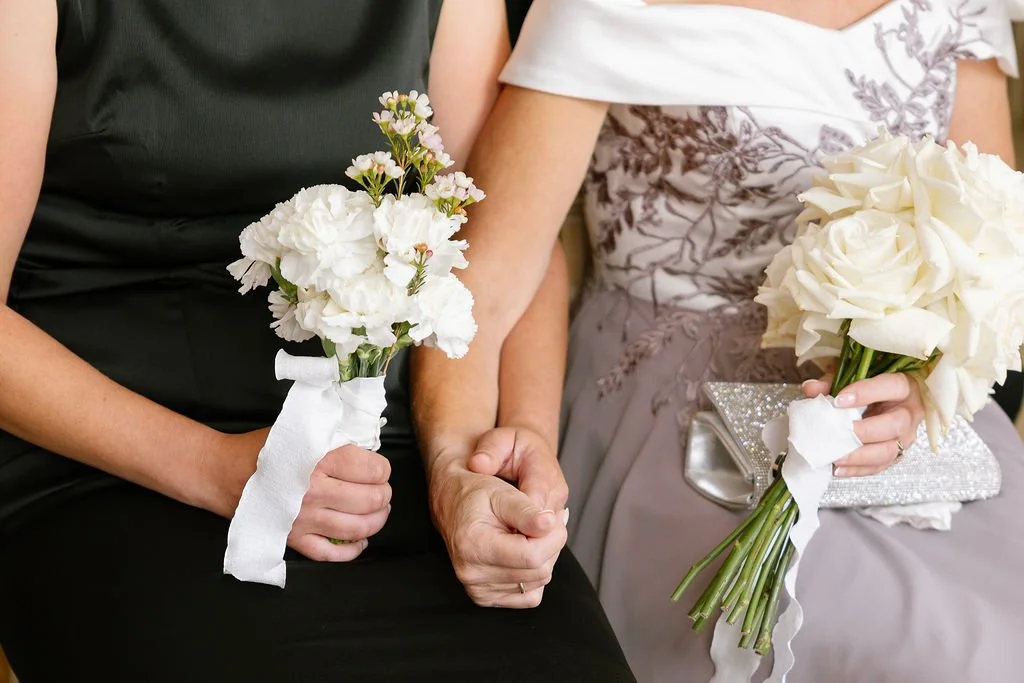 Two women sitting together, holding bouquets of white flowers, with one woman wearing a black dress and the other in a floral dress.