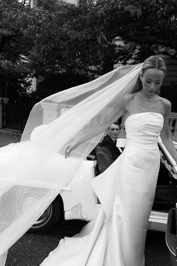 A woman in a strapless wedding dress with a long veil standing next to a vintage white car. A man in a suit is sitting inside the car, looking at her. The background features trees and a house.