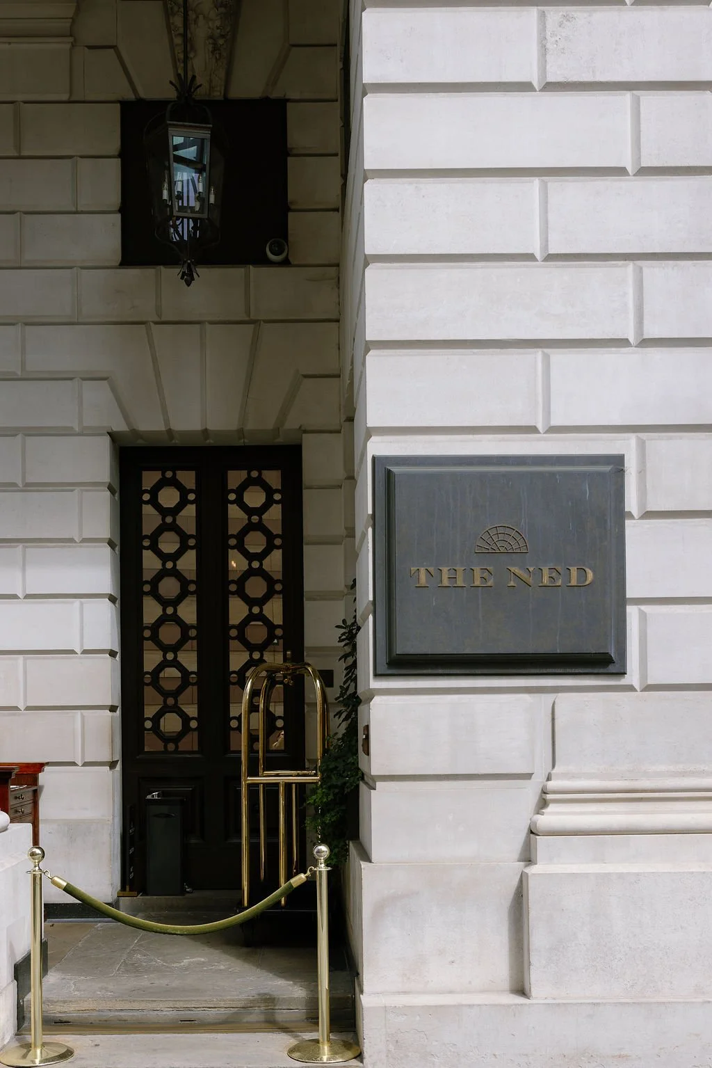 Facade of a building with a black door, a sign reading 'The Ned,' and a decorative black lantern hanging above.