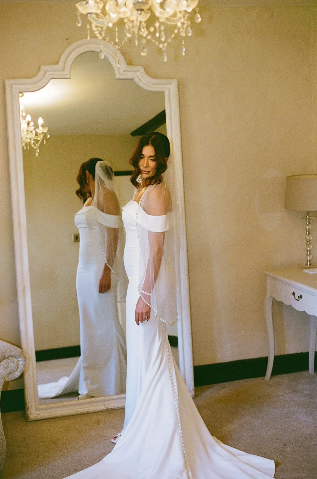 A woman in a white wedding dress with a veil is standing in front of a large mirror, smiling softly, in a room with a chandelier and a white side table.