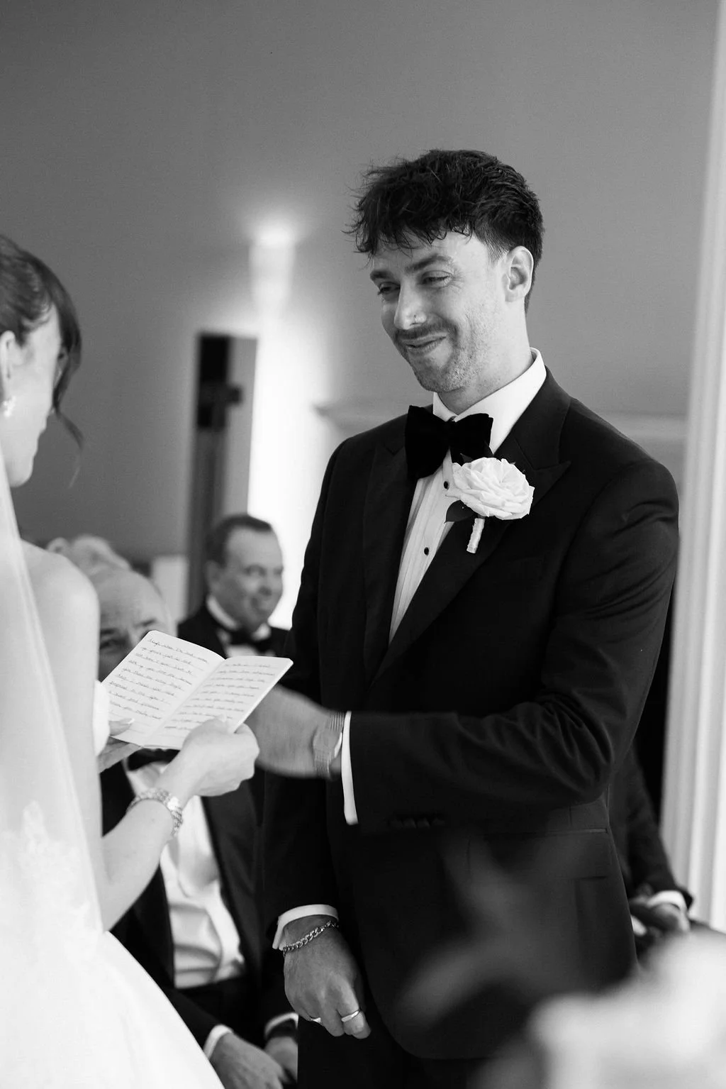 Black and white photo of a groom in a tuxedo with a white flower boutonniere, holding the hand of the bride during their wedding ceremony.