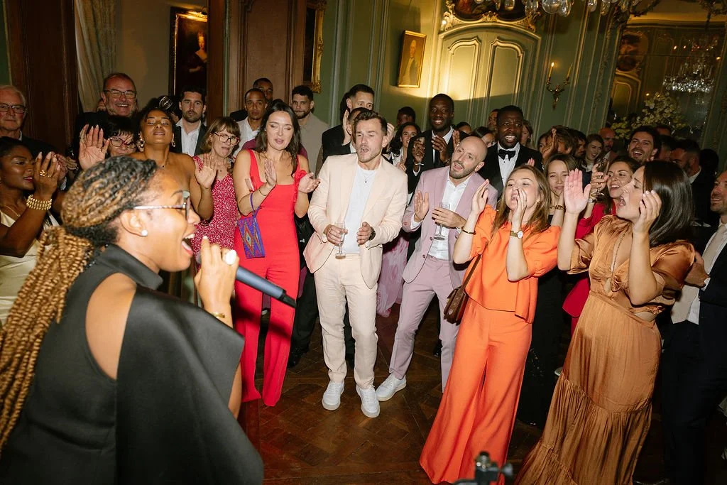 A woman with braided hair and glasses singing into a microphone in front of a lively crowd at a party or celebration in an elegant room.