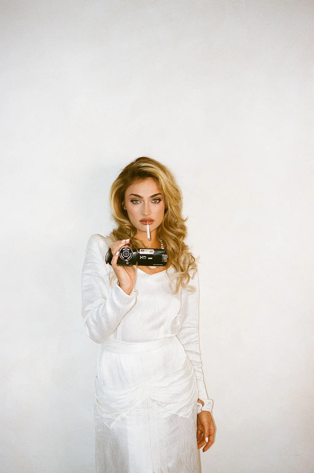 A woman with long blond hair holding a camera in one hand, with a cigarette in her mouth, wearing a white dress, standing against a plain white background.