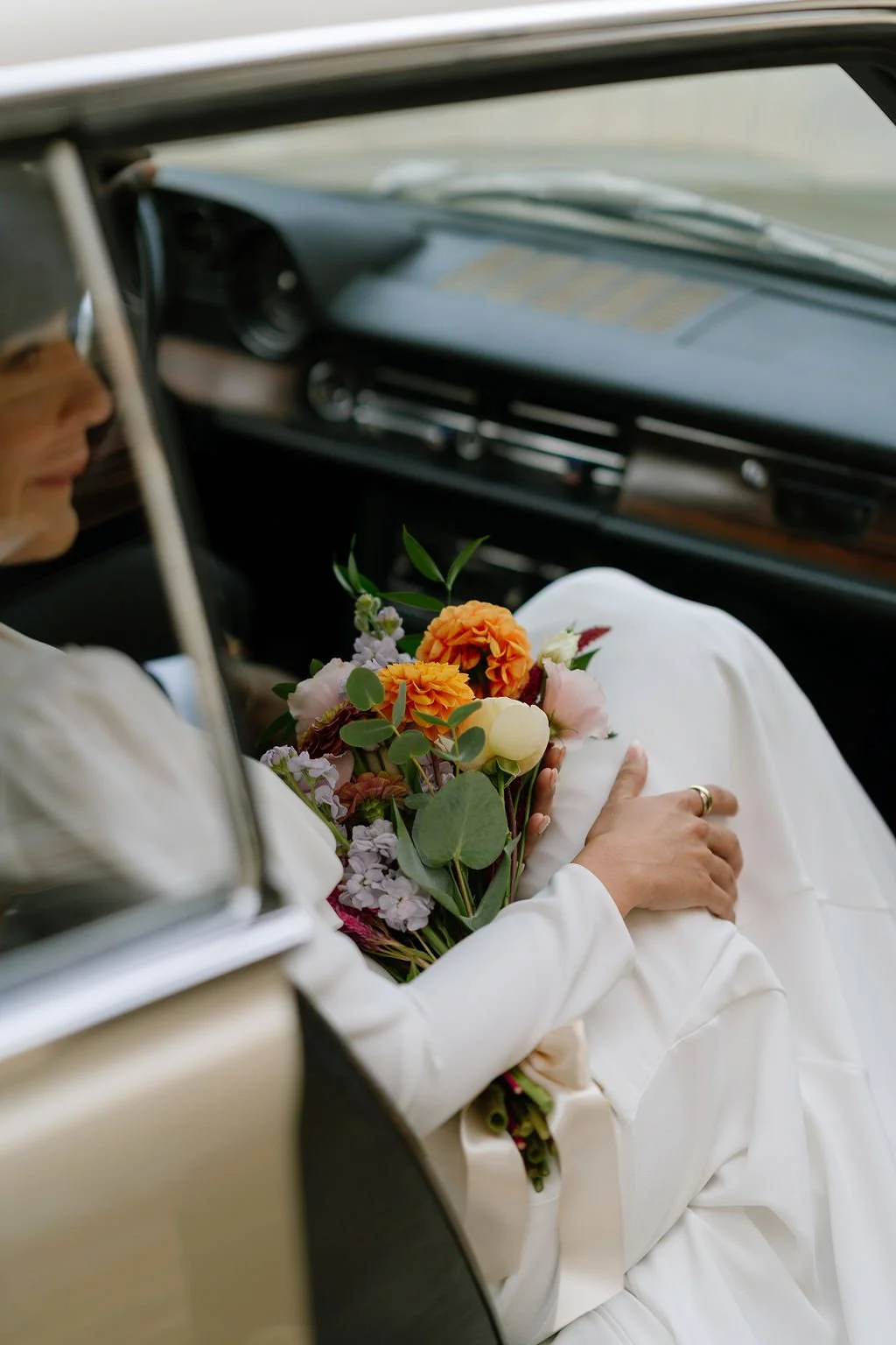 A person dressed in white holding a bouquet of colorful flowers inside a vintage car.