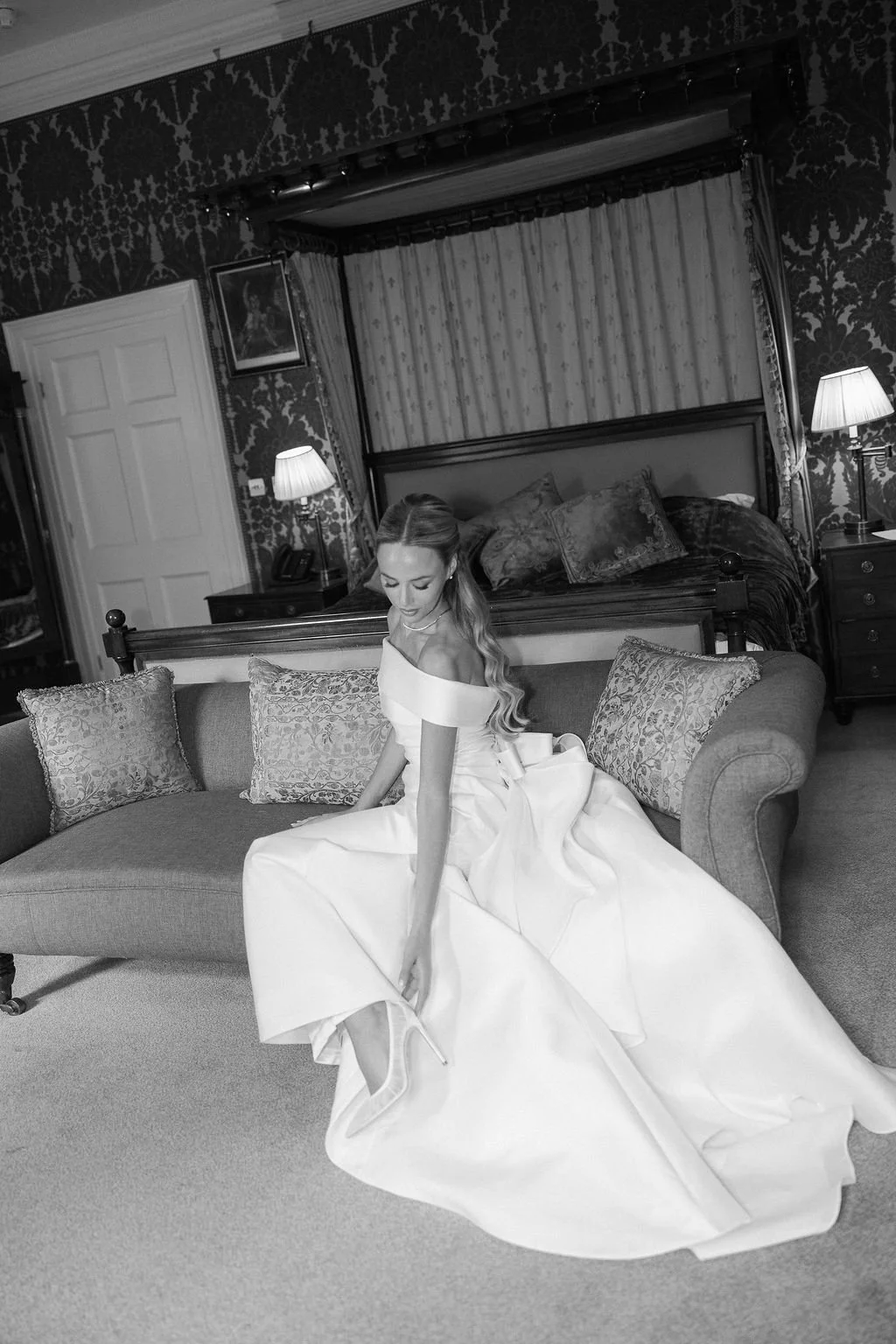 A woman in a wedding dress sitting on a sofa in a vintage-style bedroom, adjusting her high heel shoe.