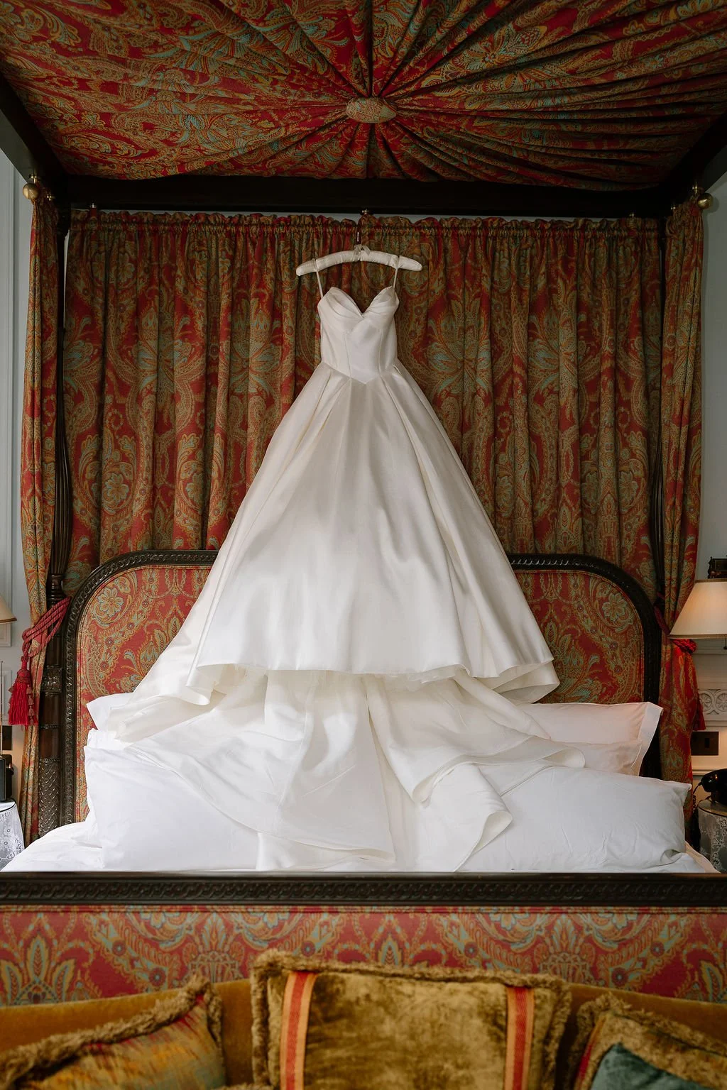 A white wedding dress hanging from a hangar on a bed with red and gold patterned curtains in the background.