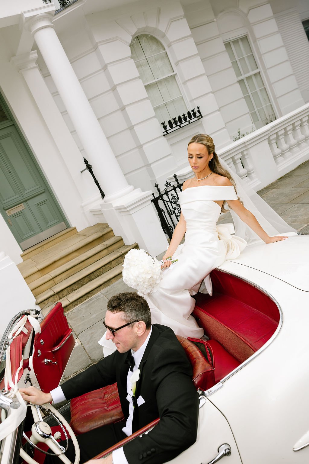 Bride in a white wedding dress sitting on the back of a vintage white convertible car with red interior, being driven by a man in a black tuxedo and sunglasses, in front of a white mansion with steps and black iron railing.