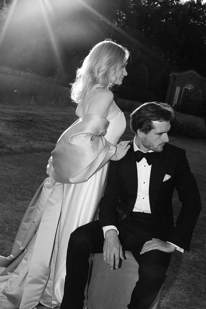 Black and white photo of a woman in an elegant dress standing beside a seated man in a tuxedo outdoors during sunset, with sunlight streaming in the background.