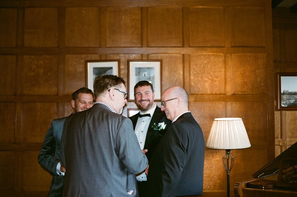 Four men dressed in tuxedos standing and talking in a wood-paneled room, smiling and laughing.