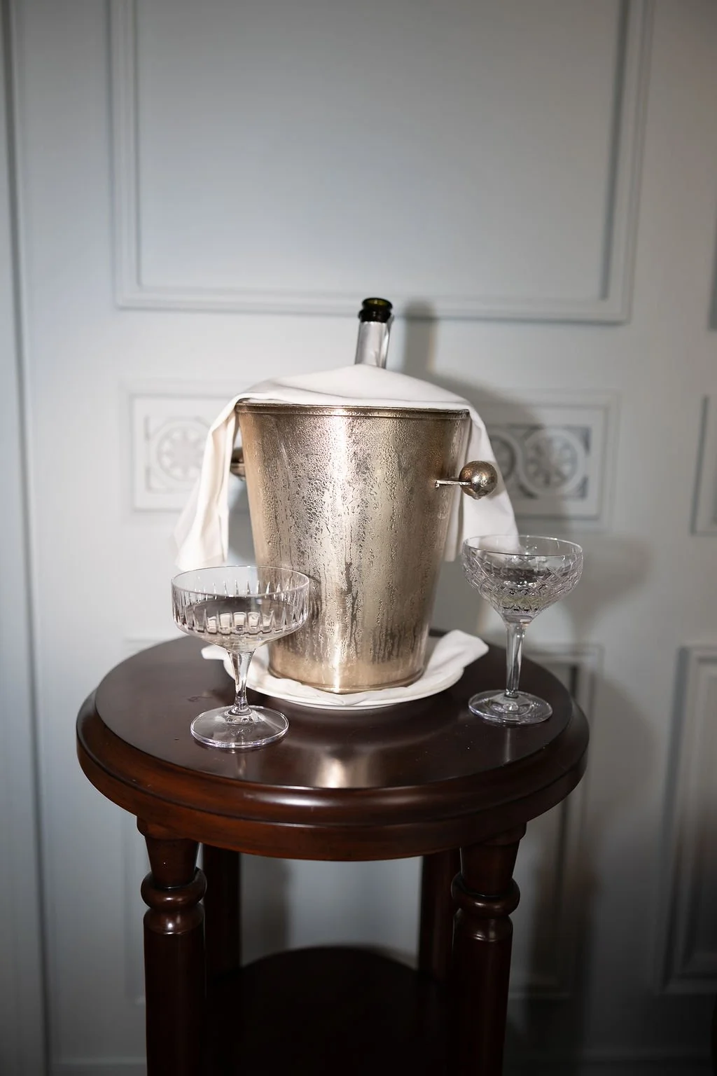 A champagne bucket with condensation holds a bottle of champagne, placed on a wooden side table with two empty glasses, against a plain wall background.