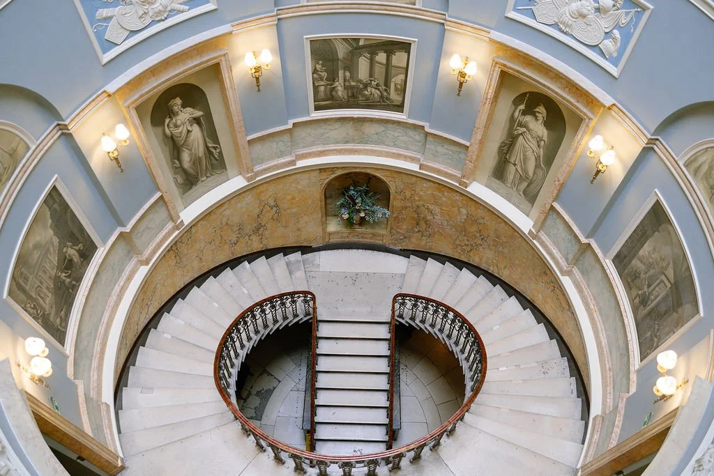 A spiral staircase viewed from above inside a building with blue walls, ornate framed black and white artwork, statues, and vintage wall sconces.