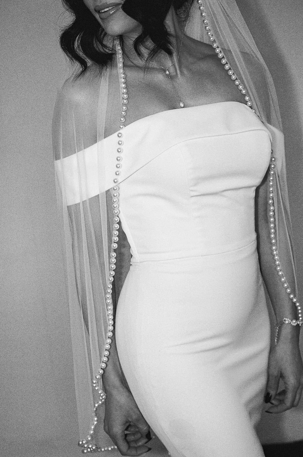 A woman in a strapless white dress with jewelry, including a pearl necklace and bracelet, standing against a plain background.