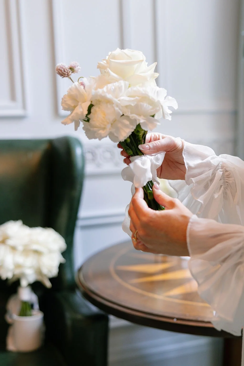 Person holding a small bouquet of white flowers, including a white rose and carnations, indoors with a green chair and wooden table in the background.