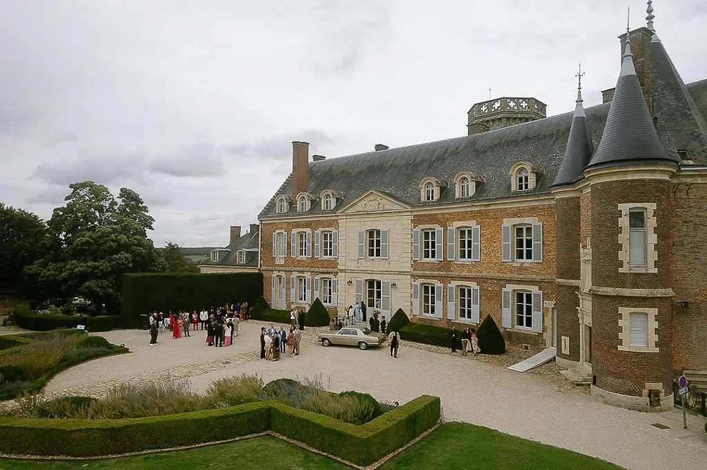 A historic chateau with a group of people gathered outside, dressed formally, on a wedding or event day. The architecture features brick and stone construction, dormer windows, and turret-style towers, set in landscaped gardens with gravel pathways.