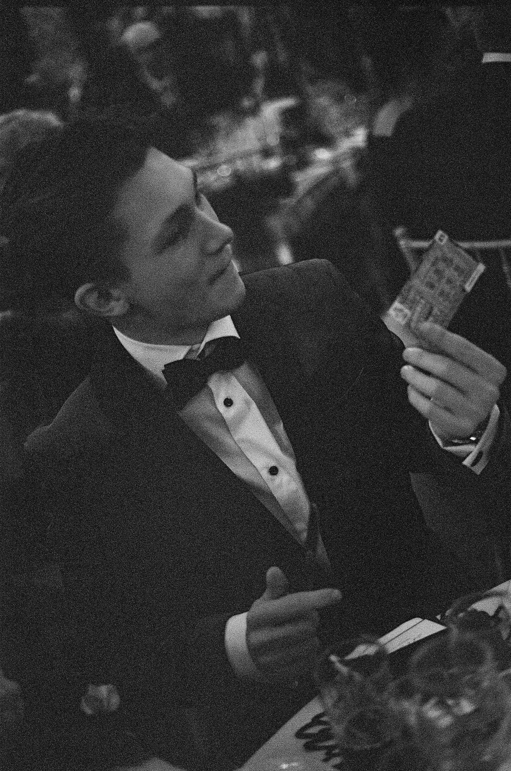 A young man in a tuxedo with a bowtie, sitting at a table, looking at his phone in a dimly lit setting.