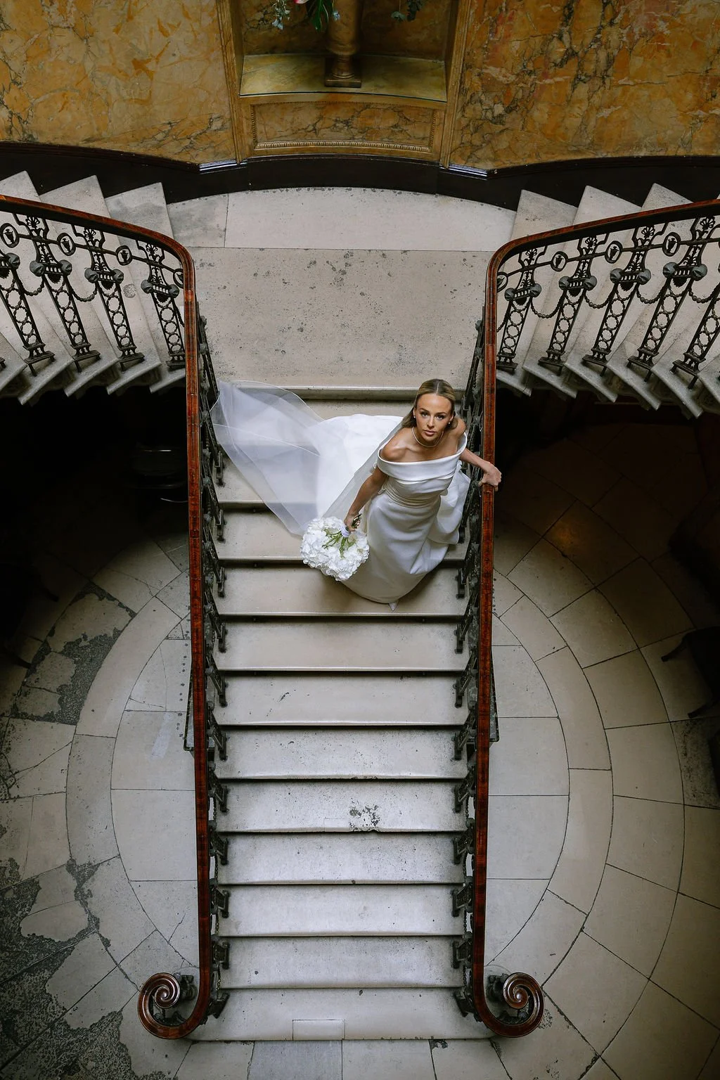 A bride in a white wedding dress holding a bouquet of white flowers, standing on a staircase viewed from above.