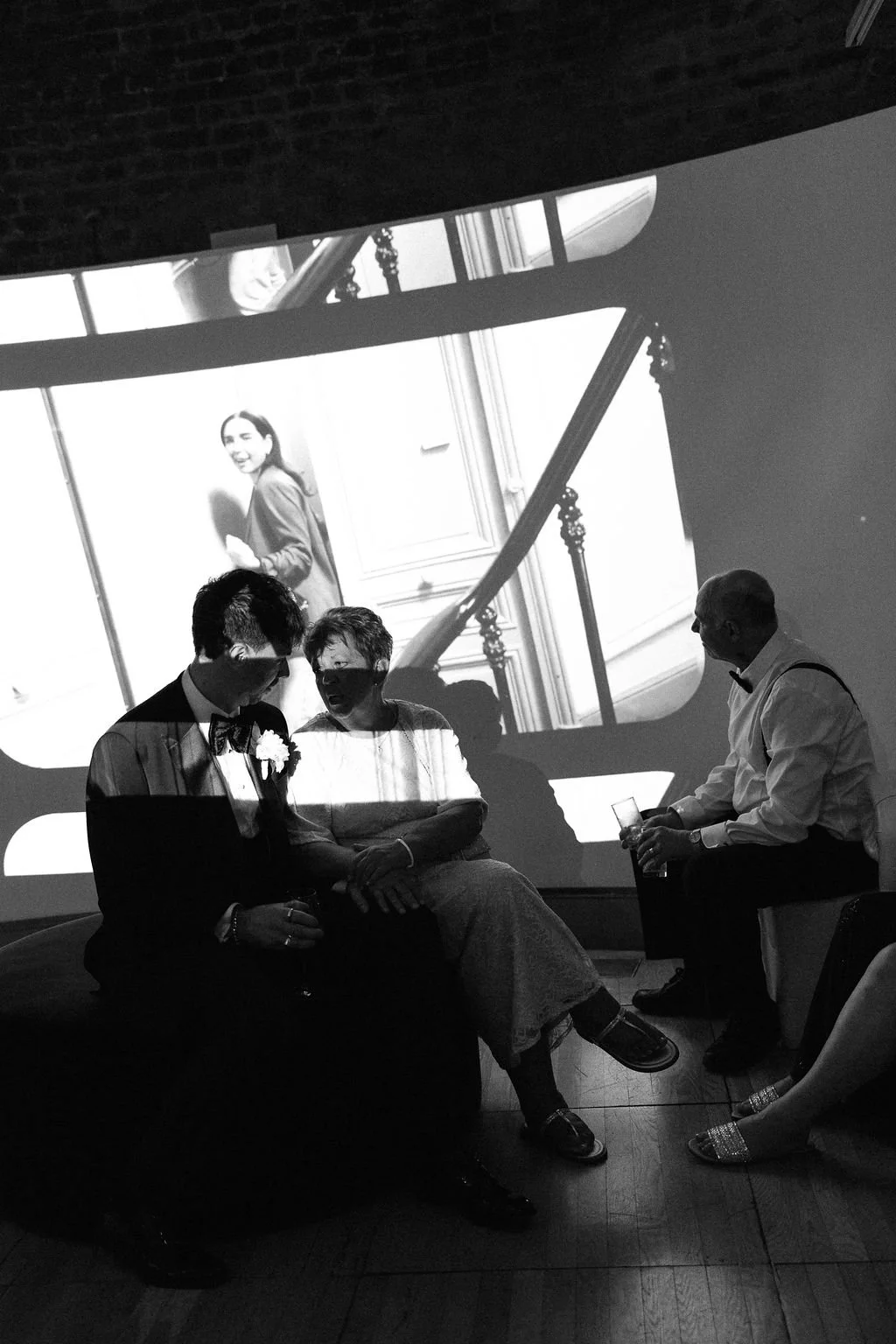 Black and white photo of three people sitting together, with a large projected image of a woman smiling on the wall behind them.