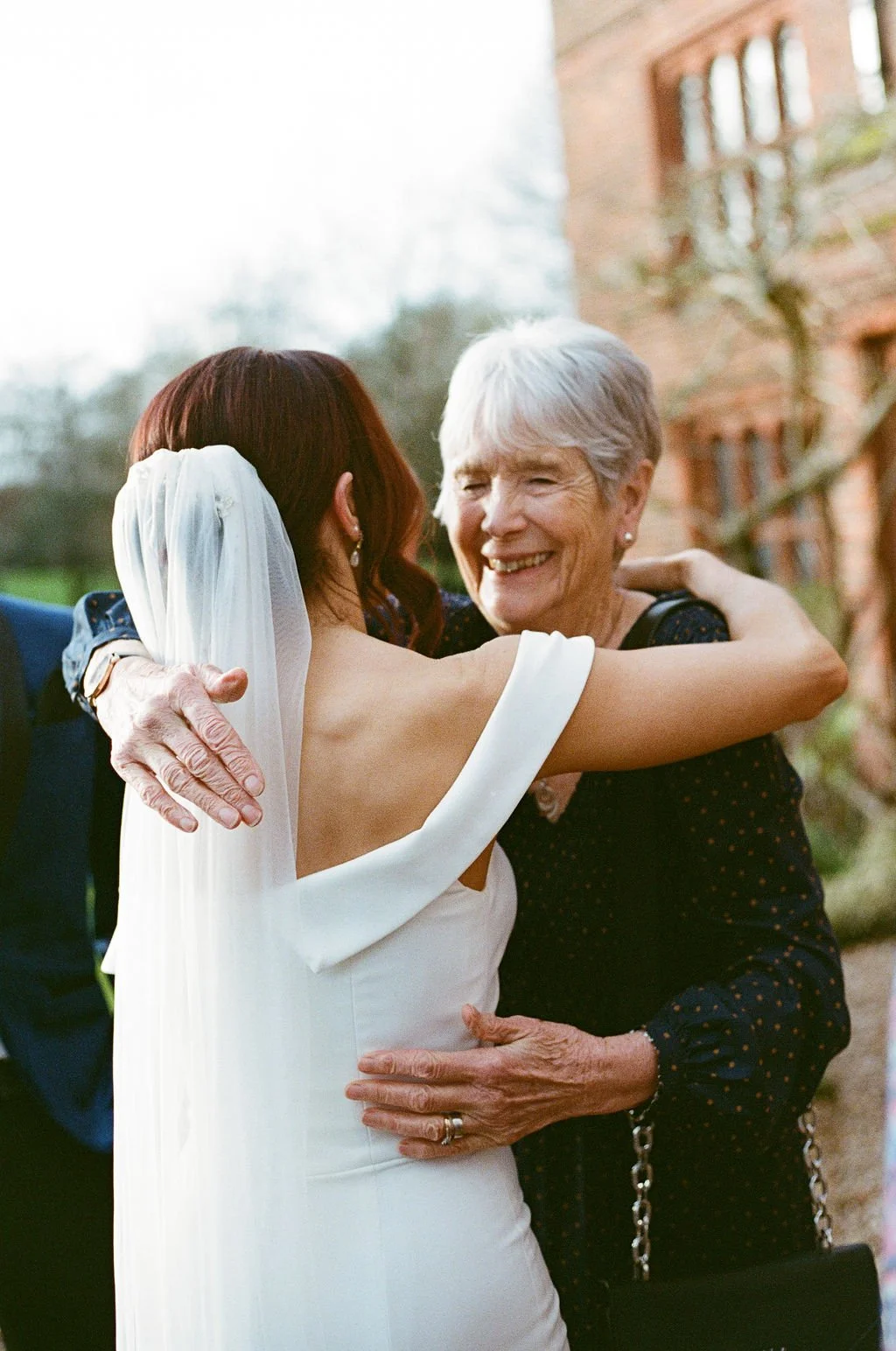 A bride in a white wedding gown and veil hugging an older woman outdoors with a brick building in the background.