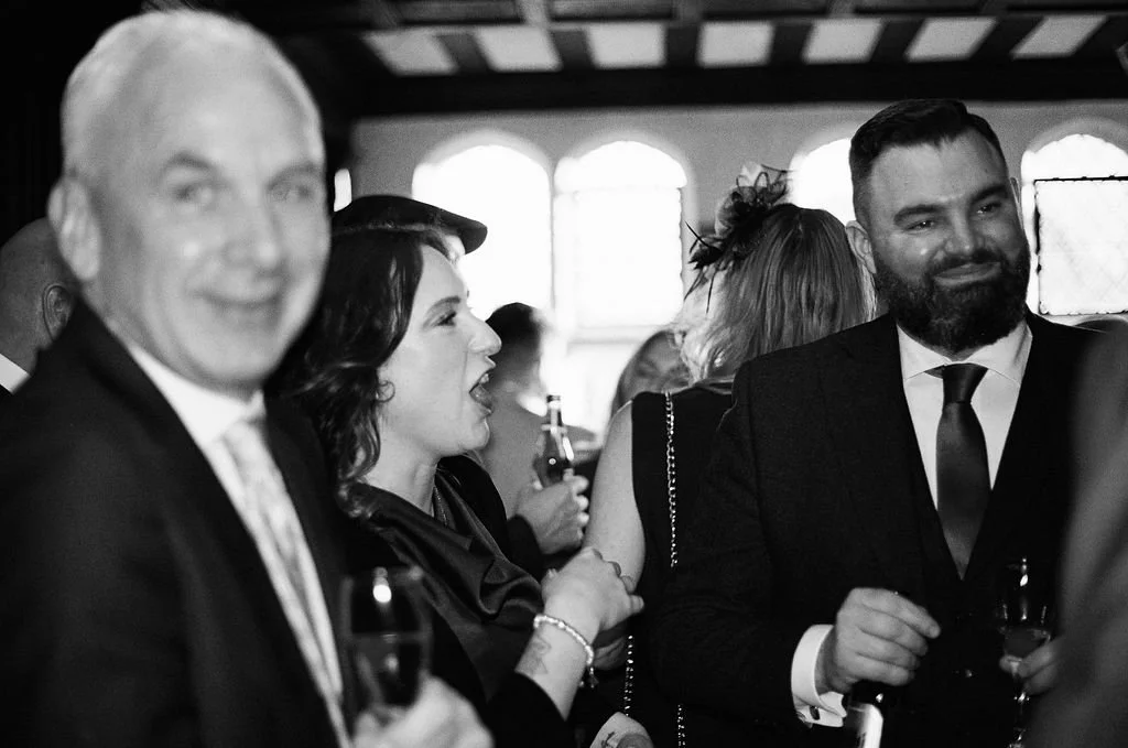 People socializing at a formal event, dressed in suits and dresses, holding drinks, inside a venue with arched windows.