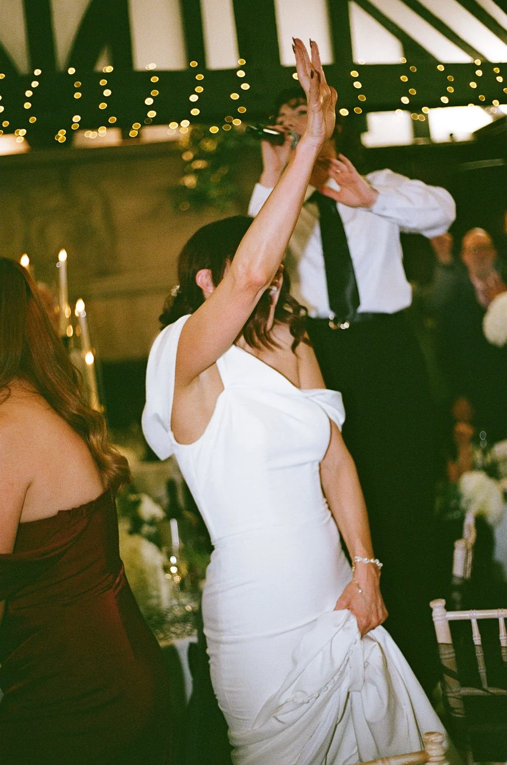 A woman in a white dress raising her arm while dancing at a celebration or wedding reception, with a man in a white shirt and black tie singing into a microphone in the background.