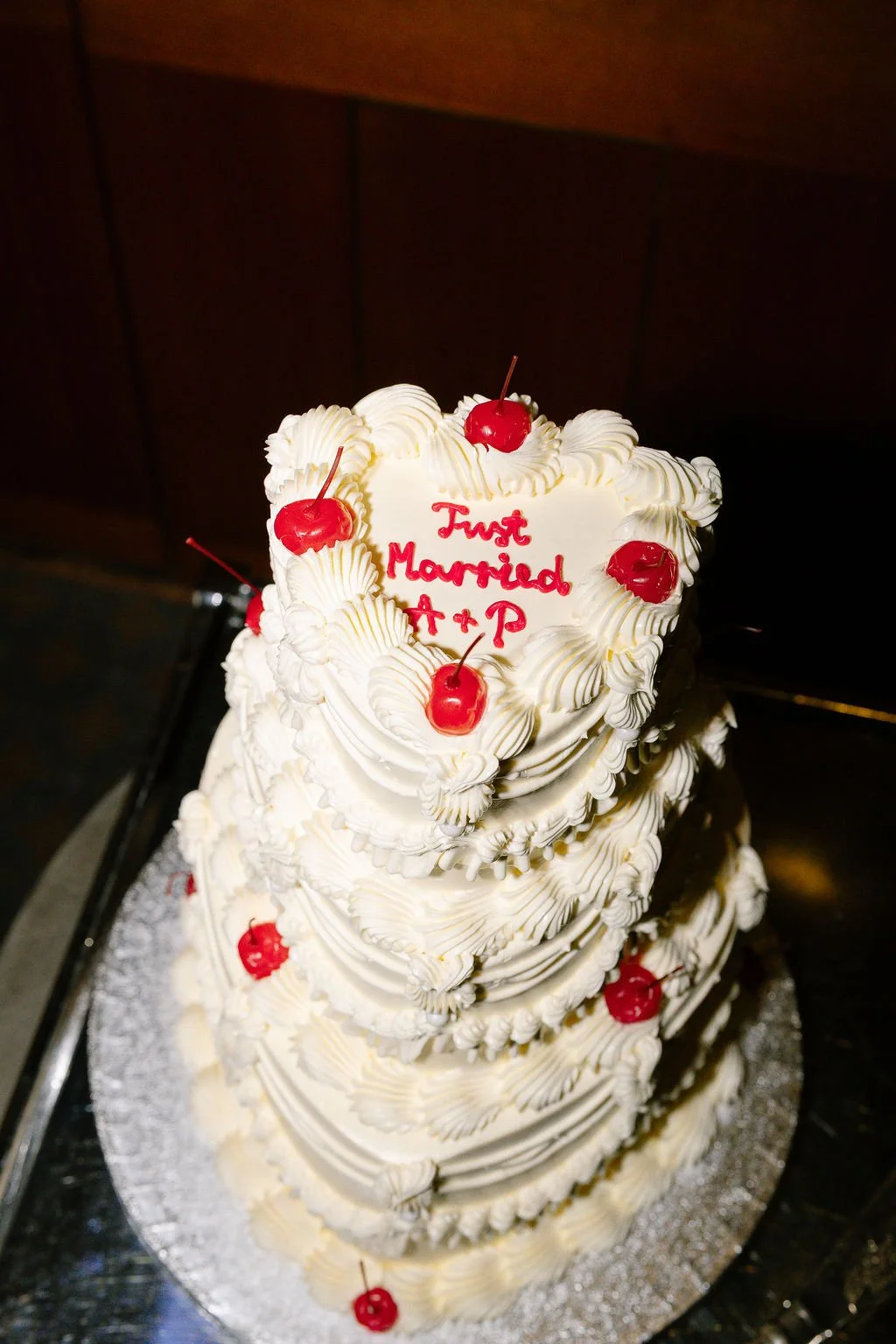 A three-tier heart-shaped wedding cake decorated with white frosting and maraschino cherries, with a message written in red icing that reads "First Married A + P."