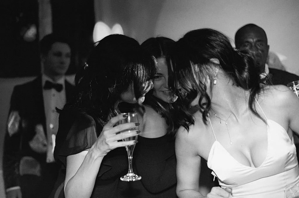 Women at a party, one holding a glass of champagne, all smiling and close together, in a black-and-white photo.