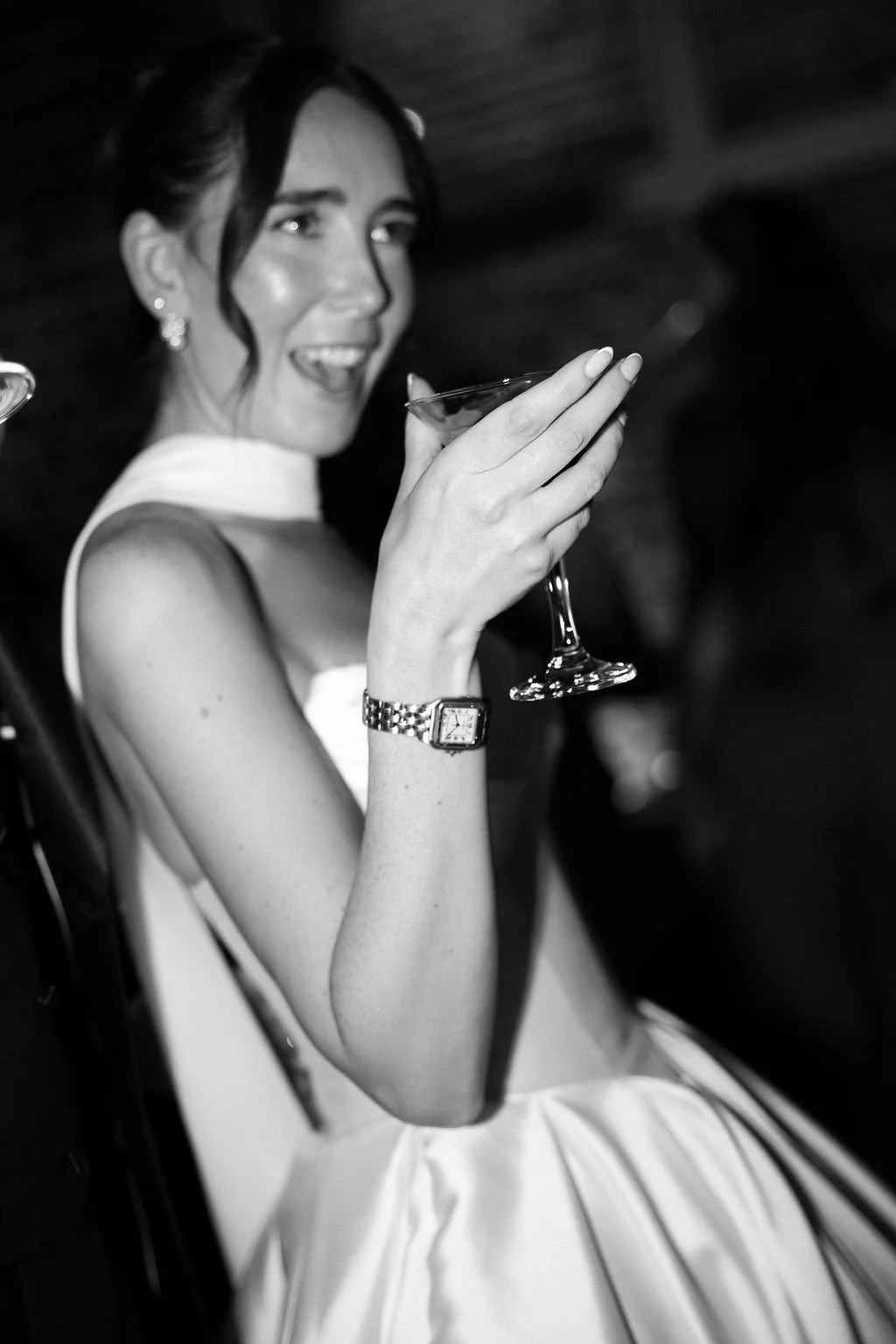 Woman in a strapless dress holding a martini glass, smiling and laughing, with dark hair styled up, wearing earrings and a watch, in black and white.