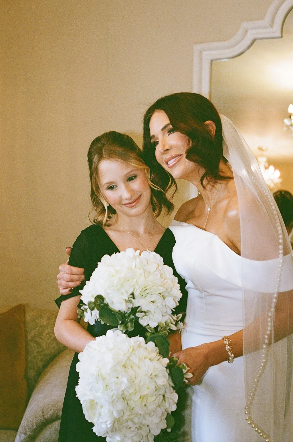 Two women, one wearing a wedding dress and veil, and the other in a black dress, standing together indoors, holding white floral bouquets, smiling.