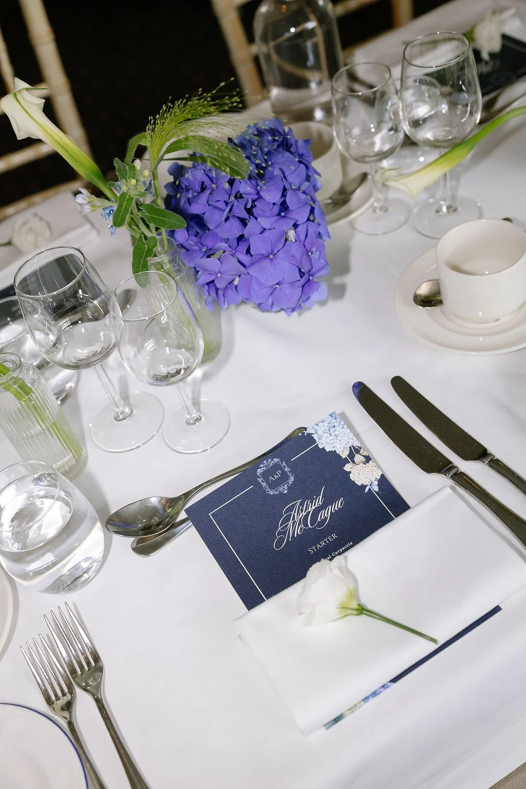 Wedding table setting with a floral centerpiece of purple hydrangeas and white flowers, glassware, silverware, a place card for Auhid McClague, and a white napkin with a white flower on top.