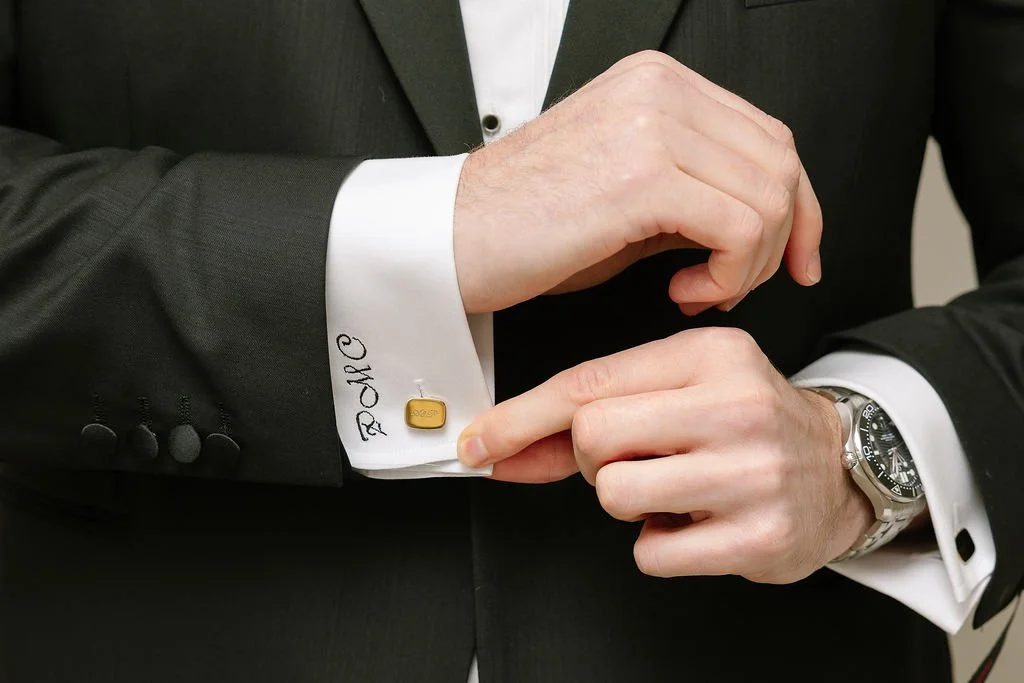 A man in a dark suit with white shirt fastening a gold cufflink on his shirt cuff.