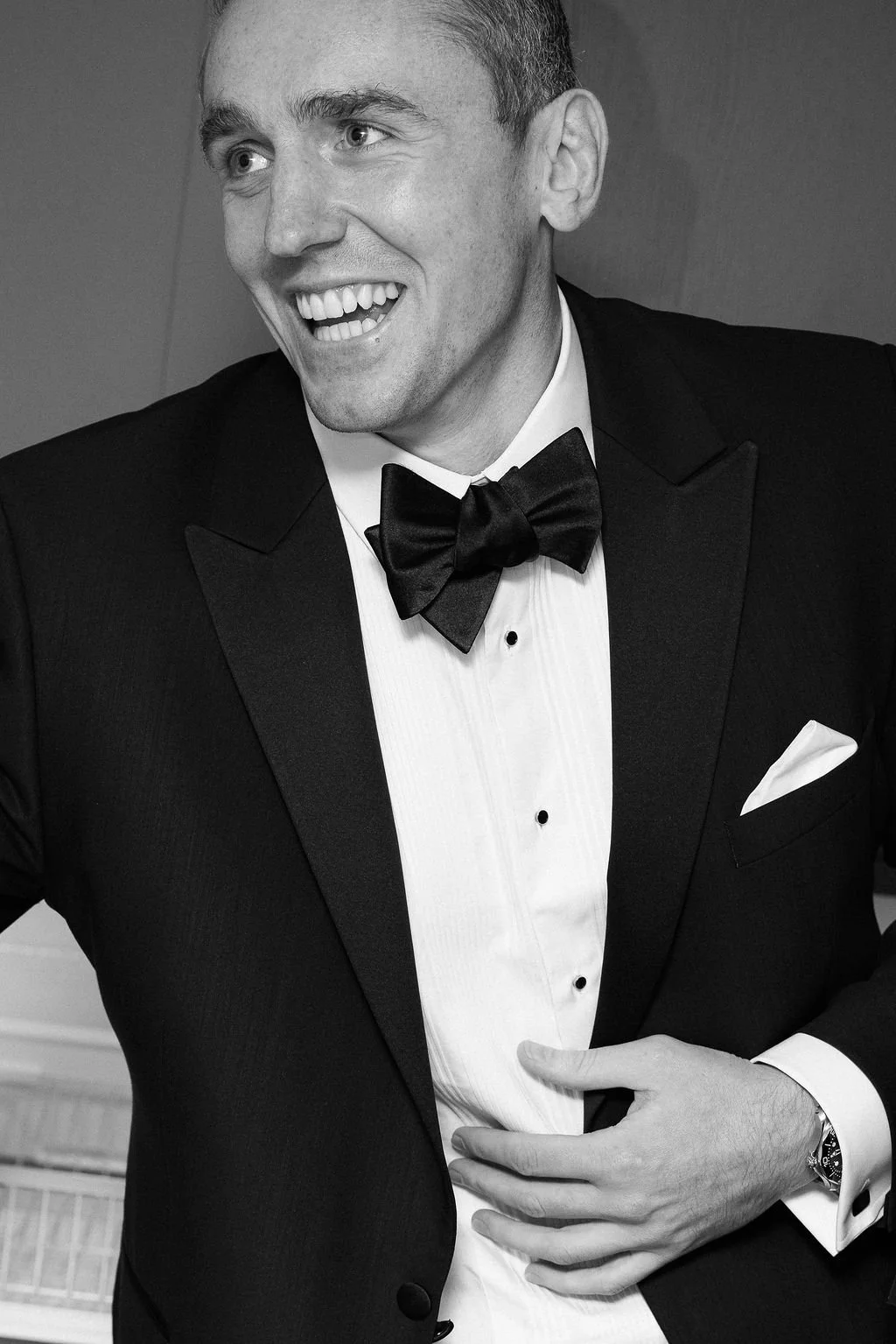 A smiling man dressed in a tuxedo with a bow tie, white dress shirt, and cufflinks, wearing a watch, and holding his stomach.